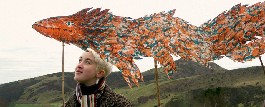 A woman stands in front of a low hill with a sculpture of a dragon.