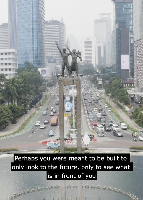 A still from Smashing Monuments. A tall concrete monument featuring two human figures, above an eight lane highway. Overlaid subtitles read: 'Perhaps you were meant to be built to only look to the future, only to see what is in front of you.'