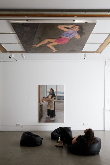 Two paintings of a woman are visible on the roof and wall of a gallery space; a figure looks at both while sat on a black bean bag.