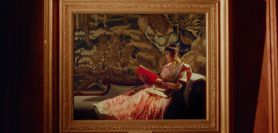 Still from art film by Aqsa Arif. A Pakistani woman lies on a chaise longue, reading a book. She is framed by a picture frame, which hangs in front of a red wall.