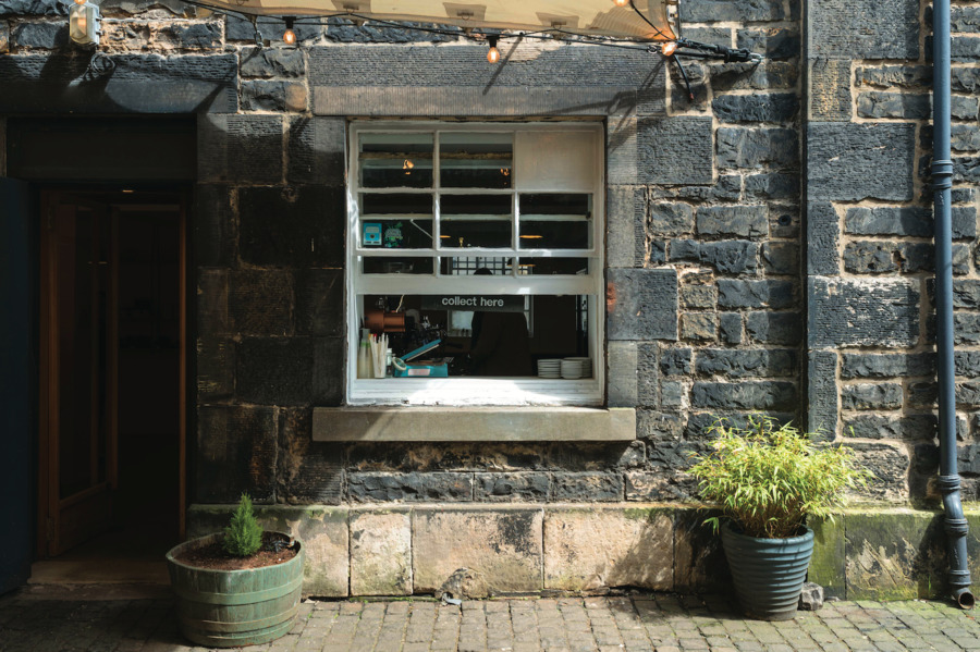 A takeaway window on the front of a stone building. There are two large potted plants, one either side of the window.