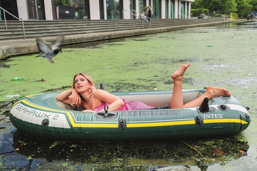 Olga Koch in a rubber dinghy on a small body of water topped with algae. A pigeon is flying into shot.