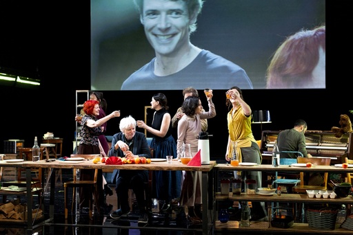 A group of actors raise glasses to toast, while one sits at a long table. Behind the group is a video screen, showing a smiling man.