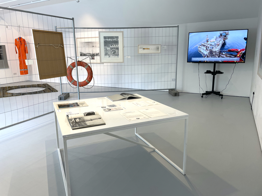 An exhibition space with a table in the centre, a screen on the right, and two sheets of fencing on the left. The exhibits appear to relate to the oil industry.