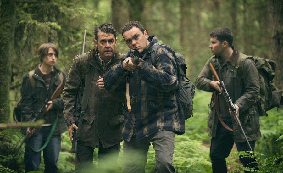 Four men stand in a forest clearing with rifles. The oldest man appears to be directing another towards a target.