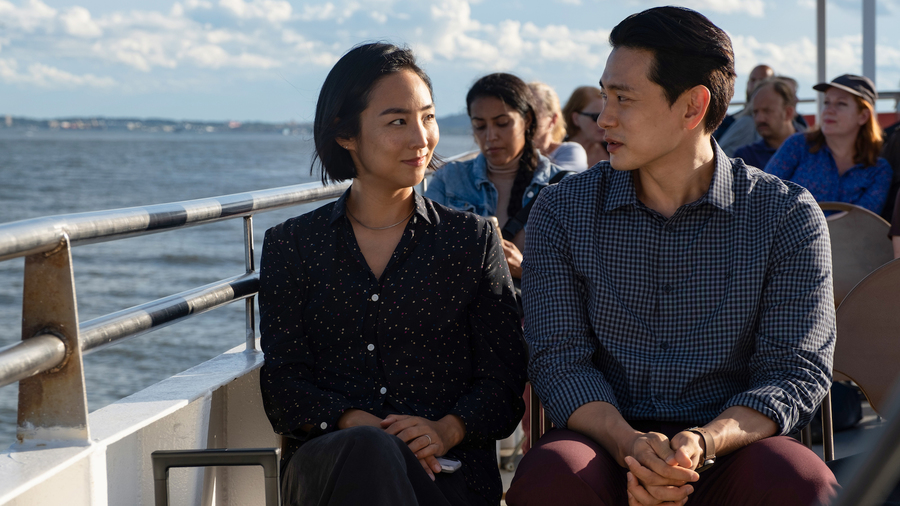 Still from Past Lives. An Asian woman and man sit side by side on the deck of a boat, looking at one another.