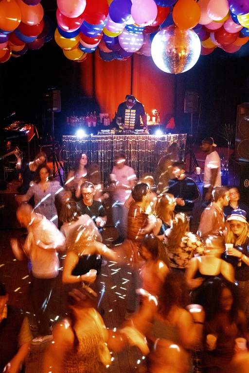 A crowd on a dancefloor; a disco ball casts lights across the room. A DJ stands behind turntables on a stage at the back of the room.