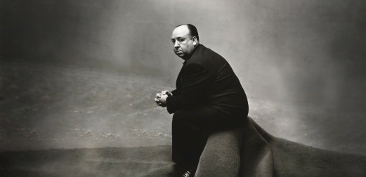 A black and white photo of Alfred Hitchcock sitting on a sparse set and looking over his shoulder