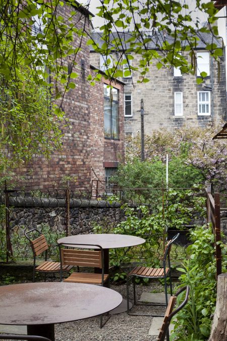 A patio seating area behind a row of tenement flats, with chairs and tables. Foliage is present all around