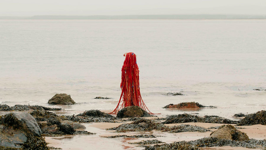 A figure with red cloth tendrils stands among some mossy rocks on a beach.