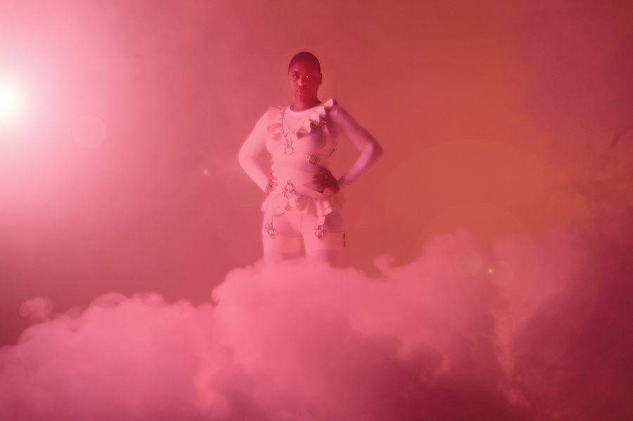 A Black woman in a pink bodysuit with frilled decorations stands in front of a pink backdrop, with a plume of pink smoke in front of her feet.