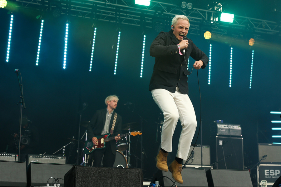 Hamish Hawk on stage at TRNSMT festival. Hamish is pictured mid-jump, singing into his microphone; he wears a dark jacket and white trousers.