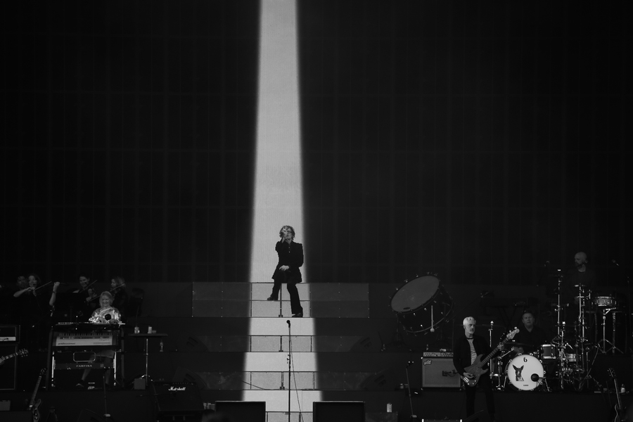 Black and white photo of Pulp on stage at TRNSMT. Jarvis Cocker sits on top of a staircase, with a spotlight shining down on him. Members of the band are visible to his left and right.