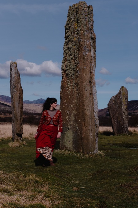 A woman in a red dress stands among a group of standing stones.