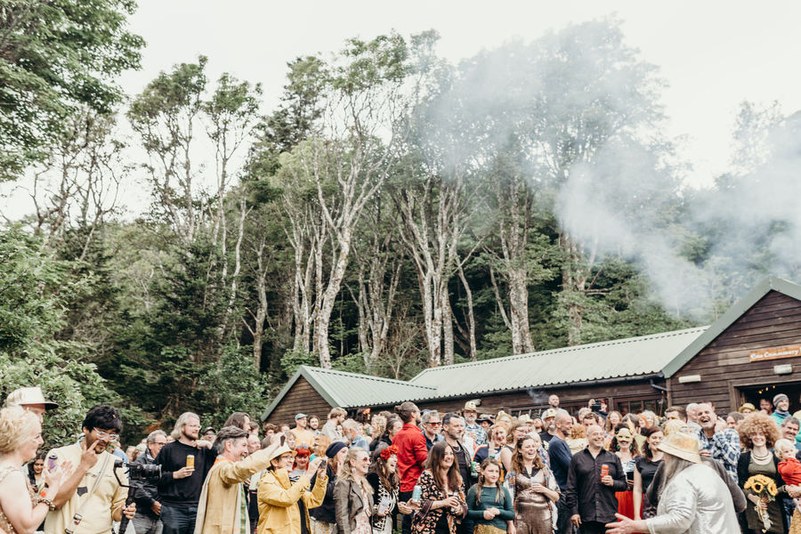 A crowd watches a performance in front of a large wooden building surrounded by trees.