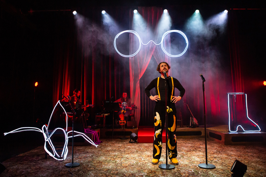 Production photo of the play Dear Billy. A man in a suit decorated with illustrations of bananas stands by a microphone, while two musicians play guitars at the back of the stage. Neon signs depicting a pair of glasses, a boot, and a plant are dotted around the stage.