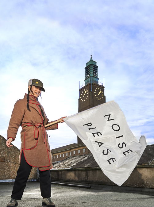 Karen Reilly of KlangHaus waves a large white flag with the words 'Noise Please' printed on it. She stands on a roof in front of a clock tower.