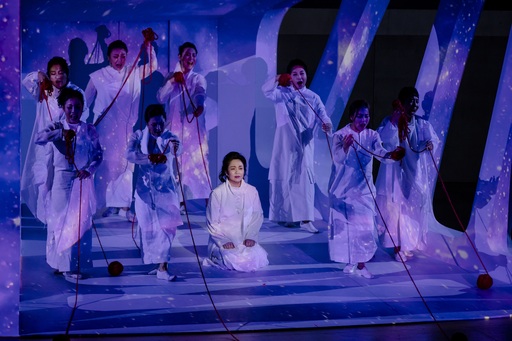 A single woman in a white gown kneels on the stage. She is surrounded by women in similar gowns, standing and pulling on reigns. The entire stage is lit in purple and blue colours.