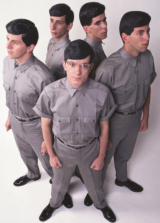 The members of Devo stand in front of a white background, each one wearing a uniform of silver shirt, silver slacks, and an overly gelled hairstyle.