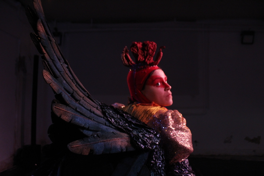 Production photo for Chicken. A woman is dressed in a chicken costume with large feathered wings and red eye make-up.