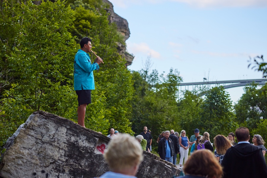 Darryl Tonemah plays a flute while standing on a large rock. A crowd has formed around him.