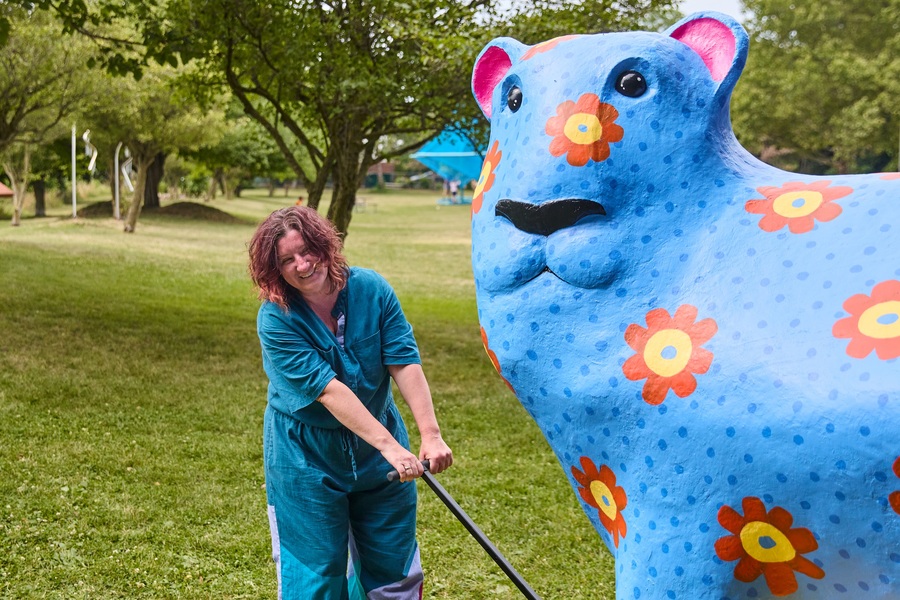 Sonia Kozlova Clark pulls a large blue sculpture of a bear-like animal across an area of parkland on a trolley.