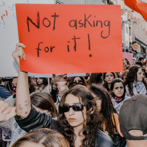 In a large crowd of people, a single person wearing sunglasses holds up a red sign, handwritten with the words "Not asking for it!"