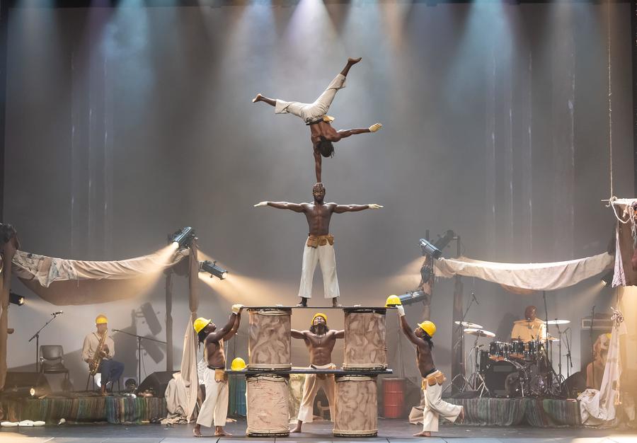A group of Black circus performers on stage. One balances on another's head, while three performers on the ground hold them in place. Two musicians can also be seen on stage.