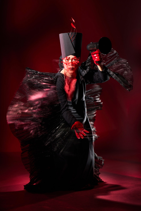 Hanna Tuulikki, dressed in a black outfit with feather-like appendages, holds out a hand while wielding a megaphone in the other.