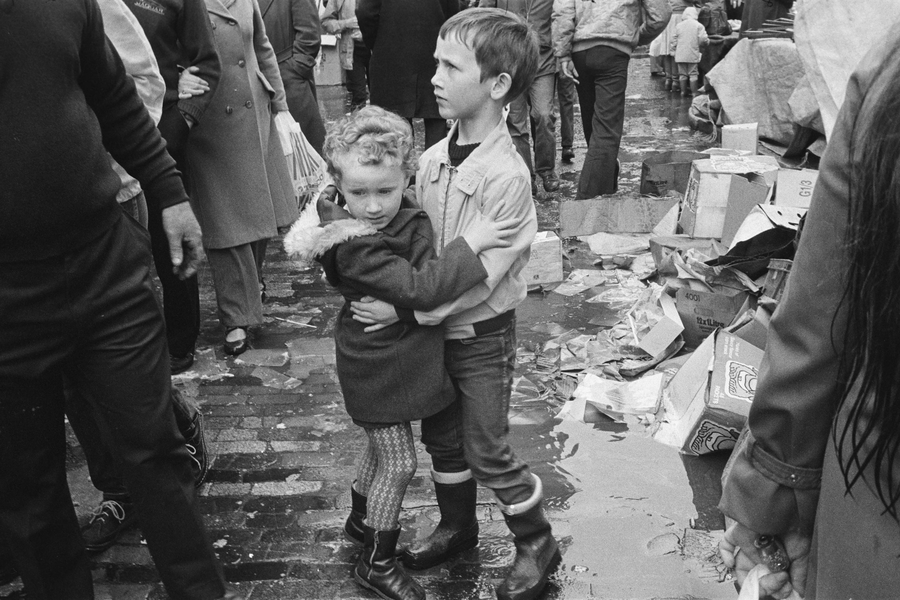 A black and white photograph of two children hugging, with a group of adults standing nearby.