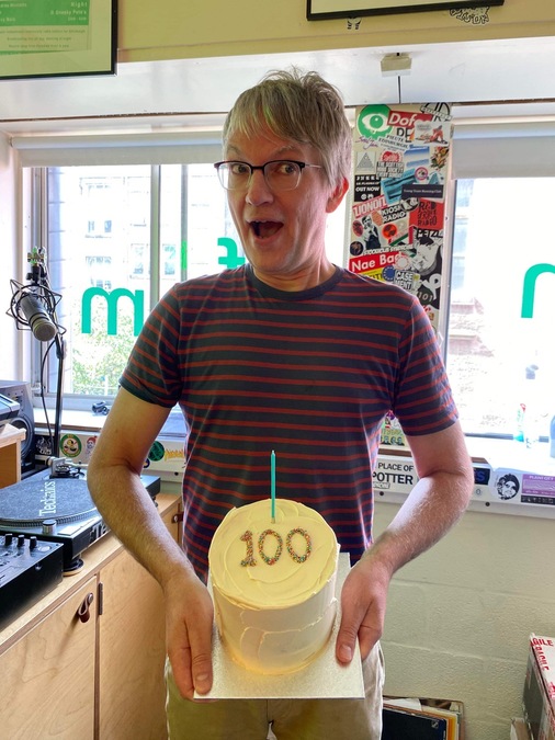 A man in a striped top stands in a radio studio, holding a cake with the number 100 written in sprinkles on the top.