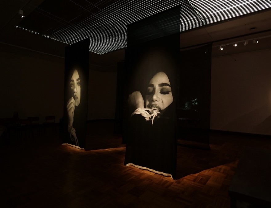 Two vertical video screens showing black and white footage of a woman. On the left she holds her hand to her face; on the right, she moves a small crab towards her mouth.