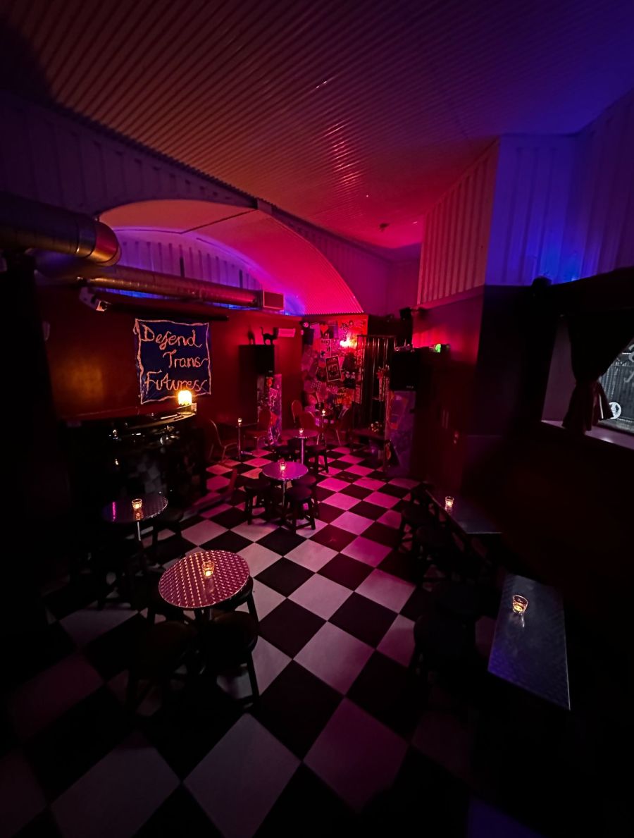 Interior photo of Bonjour. A black and white-checked floor with red walls and small tables dotted around the space; a sign reading 'Defend Trans Futures' hangs from the wall.