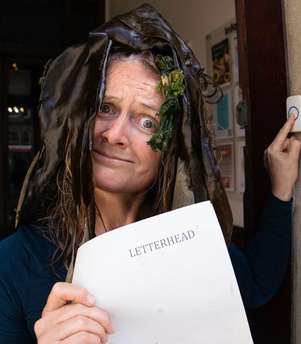 Photo of Skye Loneragan, holding a piece of paper with the word 'Letterhead' printed on it. Pieces of seaweed sit on Skye's head.