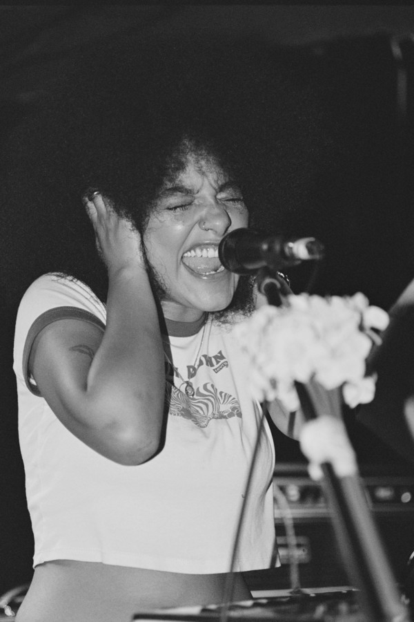A black and white photo of Lily Fontaine, singing into a microphone. A bunch of flowers are tied to the microphone stand.