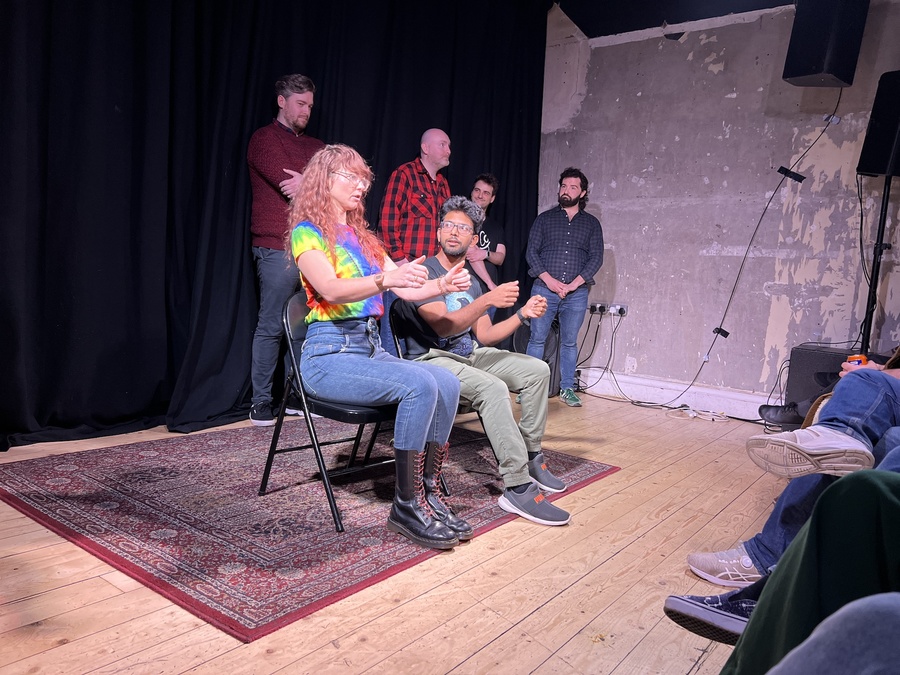 Improv comedians performing on a stage in a small room. Two sit alongside one another as if driving in a car; four performers stand behind them at the back of the stage.