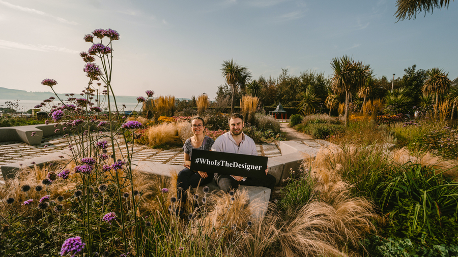 Two people sit among plants and trees at the Seabraes Viewpoint, holding a sign which reads 'WhoIsTheDesigner'
