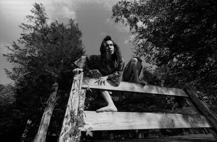 Black and white photograph of Mitski. She is climbing over a short fence in a wooded area.