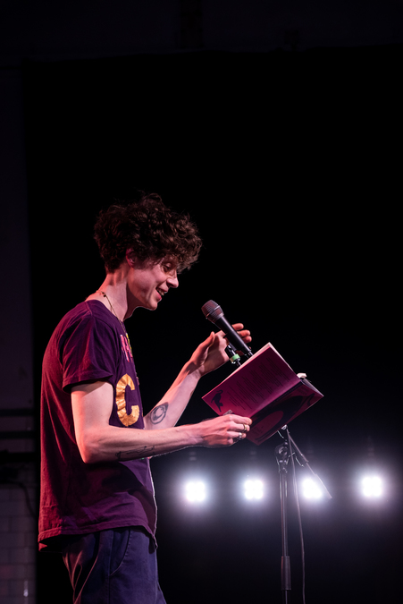 Michael Pedersen on stage, reading from a book into a microphone.