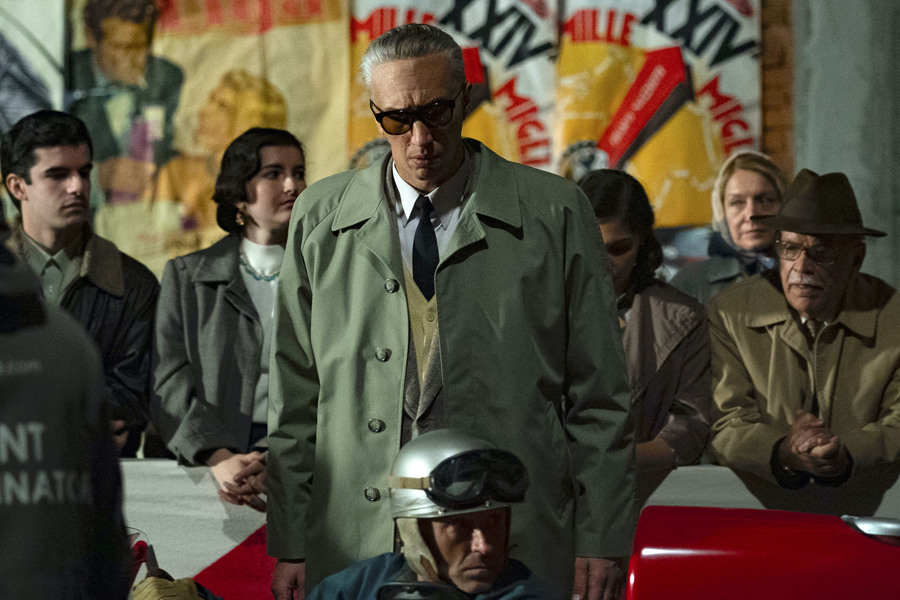 A man in a long trenchcoat and sunglasses stands by a red racecar amid a group of onlookers.