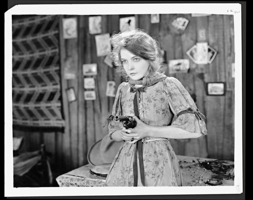 Black and white still from the film The Wind. A woman stands in a shop, holding a gun.