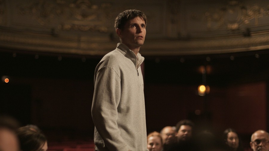 A still from Yannick. A man in a white fleece top stands up in a theatre.