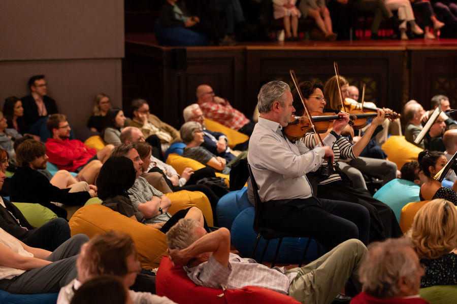 Photograph of two musicians playing violins while sat on beanbags. They are surrounded by audience members, also sat on beanbags.