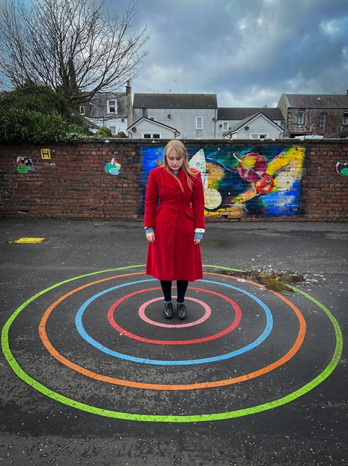 Photograph of a woman in a long red coat, standing at the centre of a series of concentric circles painted on a concrete floor in a playground.