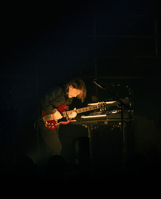 Thom Yorke playing a red electric guitar on stage. A spotlight lights Yorke and his keyboard, with the rest of the scene black.
