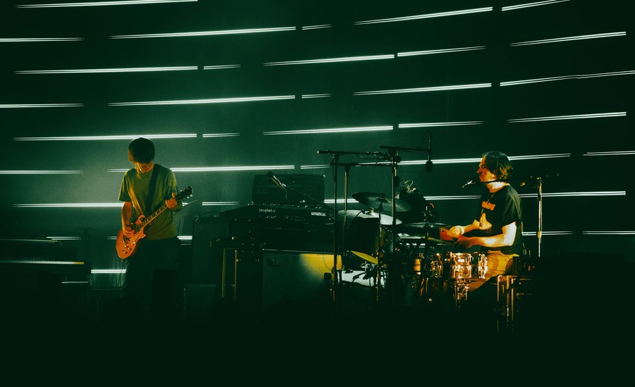 A photograph of Jonny Greenwood and Tom Skinner playing guitar and drums on stage in front of rows of thin white lights running across the stage wall.