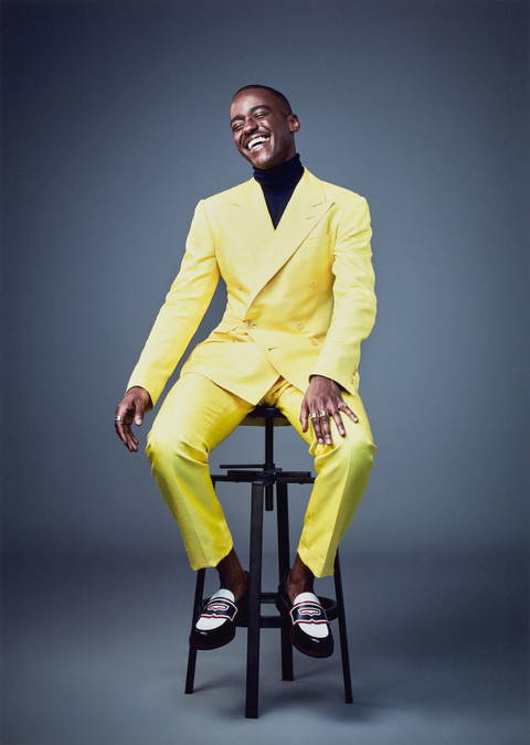 Portrait photo of Ncuti Gatwa, sitting on a tall stool wearing a bright yellow suit.