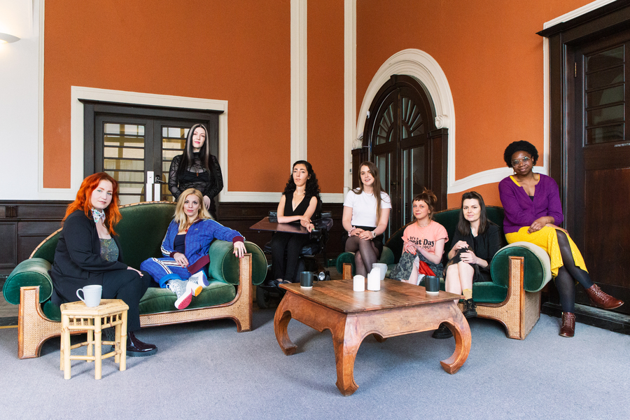 Members of the Hen Hoose musicians collective sit on a variety of chairs and sofas in an orange-walled room.