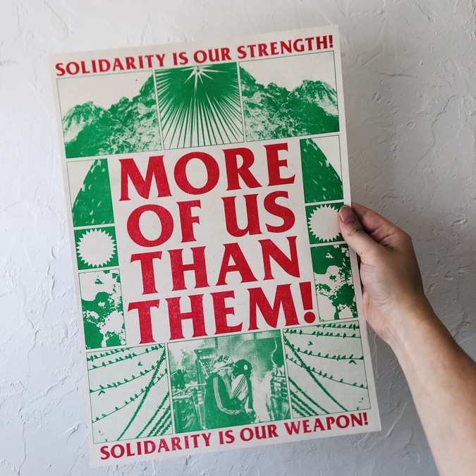 Photo of an arm holding a risograph print. Red text on the print reads 'Solidarity is our strength; More of us than them! Solidarity is our weapon!' The print also include a series of green illustrations and images.