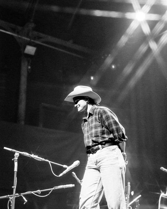 Black and white photo of Adrianne Lenker, wearing a plaid shirt, jeans, and a cowboy hat.